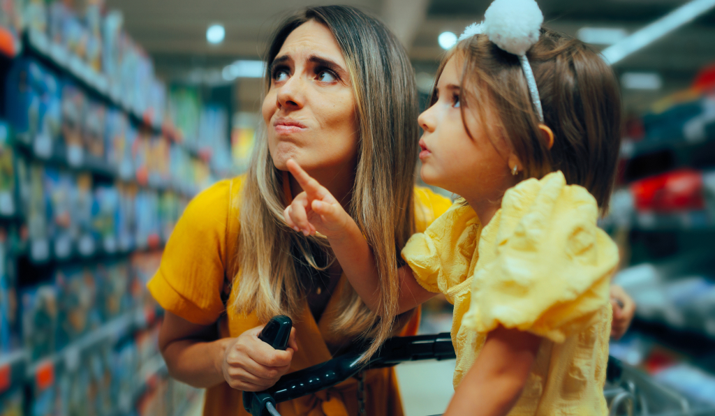 Mother and young daughter wearing yellow dresses stand in a supermarket aisle; the daughter sits in the shopping cart and points ahead while the mother looks up thoughtfully, gripping the cart handle.